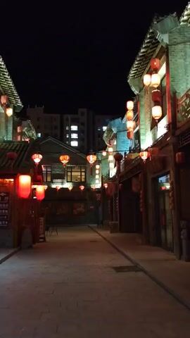 The video shows a narrow street at night, lined with traditional brick buildings adorned with red lanterns. The lanterns are illuminated, casting a warm glow on the cobblestone street. Some buildings appear to be shops or restaurants, with signs visible in Chinese characters. A few windows show internal lighting. In the background, a modern, multi-story building with lit windows can be seen, contrasting with the older architecture. The sky is dark and clear. There are no people visible in the video. The camera pans slowly down the street, offering a view of the illuminated lanterns and architectural details.