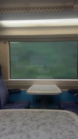A man is visible through a window on a train. He is seated at a table and holding a dark object in his right hand, which he appears to be looking at. He is wearing sunglasses and a light-colored shirt. The train car interior is visible, with blue seats and white tables. The lighting is artificial and the overall scene is dimly lit.