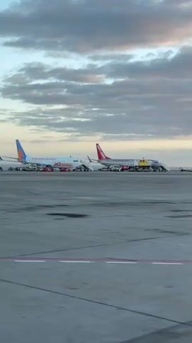 The video shows two parked airplanes on an airport tarmac under a cloudy sky. The planes are Jet2holidays and Jet2.com, and appear to be undergoing maintenance or pre-flight preparations, surrounded by ground support equipment like baggage carts and service vehicles. The tarmac is concrete, with painted lines and markings visible. There is no active movement of the planes or other vehicles.