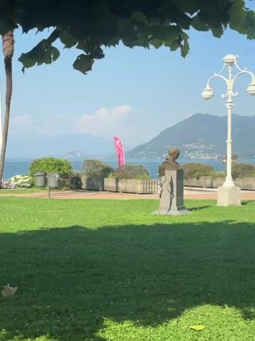 This media file captures a picturesque waterside park scene in Binda, Italy. The time of day appears to be late morning or early afternoon, indicated by the bright sunlight casting distinct shadows. The weather is clear and sunny, with a bright blue sky and a few wispy clouds. Lush green grass dominates the foreground, with trees and flowering bushes bordering a paved walkway.

The central focus of the scene is a bronze bust on a stone pedestal, likely a monument to a notable figure. To the right of the bust, a decorative white lamppost with three lights stands tall. In the mid-ground, a body of water, presumably a lake or sea, stretches towards distant, hazy mountains. A vibrant pink flag is planted near the water's edge, and a few boats can be seen on the water in the distance.

A woman wearing a maroon dress and carrying a backpack walks along the paved path, adding a human element to the serene landscape. Palm trees and other deciduous trees provide shade and visual interest. The overall mood is tranquil and inviting, suggesting a pleasant day for a stroll in the park. No visible text is present in the media file.