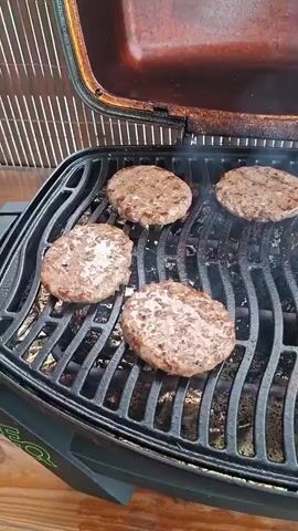 The video shows several hamburger patties cooking on a grill. The grill is black with wavy grates, and some smoke is rising from the patties. The patties are round and appear to be made of ground meat. They are arranged in a grid pattern on the grill, with some closer to the viewer than others. The grill lid is open, revealing the cooking surface and the patties. The background is slightly out of focus but appears to be a wooden surface or deck. The lighting suggests it is daytime. There are no people visible in the video. The scene is focused on the food and the cooking process. The patties are browned on the sides and appear to be cooking evenly. The overall impression is that of a casual outdoor cooking scene, likely a backyard barbecue or gathering.
