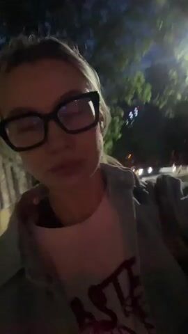 A young woman is recording a selfie video outside at night. She is wearing glasses, a pink t-shirt with a design, and a denim jacket. She touches her hair and adjusts the camera angle slightly. The background shows a street with trees, streetlights, and parked cars. The video is somewhat shaky.