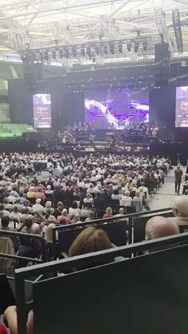 A wide shot of an indoor concert venue. A large stage is set up at the far end of a large, tiered seating area filled with an audience. The stage is lit with bright lights and features a large screen displaying abstract purple and white visuals. An orchestra, dressed in formal attire, is performing on stage, accompanied by what appears to be a conductor. The audience members are seated and facing the stage. The overall atmosphere suggests a formal musical performance.