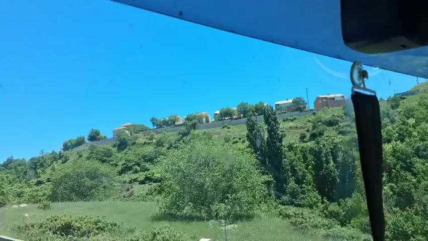 The camera is positioned inside a vehicle, looking out through the windshield. The sky is bright blue and clear. A hill rises in front of the vehicle, covered in green trees and shrubs. At the top of the hill, a row of modern-looking houses with light-colored roofs is visible. The word "TOLJA" is displayed in large letters on the hillside, near the houses. A rearview mirror and a lanyard hang from the top of the windshield, slightly obscuring the view.