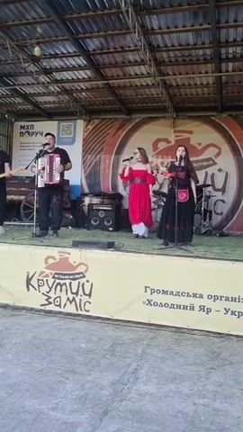 A band performs on an outdoor stage with a roof. A male accordion player, two female singers, a male guitarist, and a drummer are on stage. The accordion player is in the center-left, the two female singers are in the center, and the guitarist and drummer are on the left and right, respectively. The singers are wearing dresses, one red and one black. The stage is decorated with banners, including one that reads "КРУТИЙ ЗАМІС" in yellow and black. The ground in front of the stage is concrete.