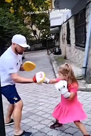 The video depicts a man and a young girl engaged in a boxing training session outdoors. The man, wearing a white t-shirt, blue shorts, and a white baseball cap, is holding boxing pads, one of which appears to be red and white. The girl, dressed in a bright pink dress and white boxing gloves, is actively throwing punches at the pads.

The setting appears to be a paved outdoor area, possibly a driveway or courtyard, with a white SUV parked in the background. Trees and foliage are visible, suggesting a daytime setting, likely warm weather given the attire. The architecture in the background includes brickwork and fencing, hinting at a residential or possibly a commercial area. The overall mood seems to be one of active training and mentorship, with the man guiding the girl's movements.

Visible text overlays in the top portion of the frame, in what appears to be Ukrainian, read "Слідкування" and "Для тебе," which translate to "Following" and "For you," respectively. These are likely captions or social media indicators. The image quality is somewhat grainy and appears to be a still frame from a video.