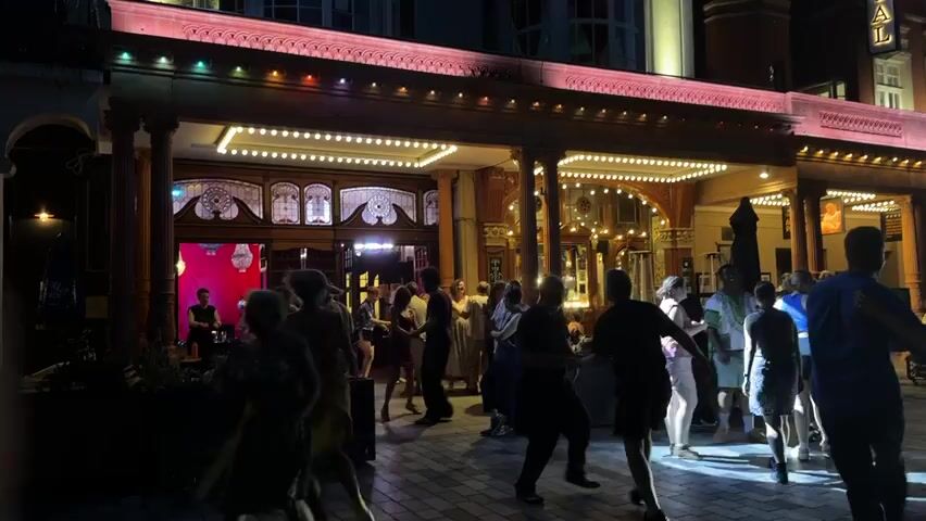 The video shows a group of people dancing outdoors in front of a building at night. The building appears to be a theater or a similar venue, with a grand entrance featuring columns, decorative lights, and a red awning. The dancers are a mix of ages and genders, dressed in casual and semi-formal attire. Some are dancing in pairs, while others appear to be dancing individually or in small groups. The lighting is dim, with the building's lights and some spotlights illuminating the scene. The ground is paved with brick or stone. The overall atmosphere seems festive and lively.