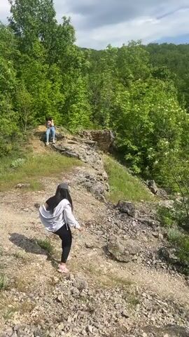 The video starts at a low angle looking up a rocky, gravelly slope. A woman in a white jacket, dark pants, and pink shoes is standing on the slope. Another woman is sitting further up the slope. The camera pans upward to show a vista of green trees, a grassy clearing, and a mountain range in the distance under a cloudy sky.