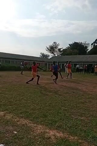 This video captures a group of young people engaged in a lively game of netball on an outdoor field in Ngenyilel ward, Kenya. The scene appears to be set during daylight hours, with a cloudy sky suggesting it might be mid-afternoon or early evening. The players are dressed in athletic attire, with some wearing red and yellow bibs over their outfits, indicating different teams. They are actively participating in the game, with one player in the foreground skillfully dribbling a basketball. The background reveals a row of buildings, possibly school facilities, and a scattering of trees, characteristic of a rural or semi-urban setting in Kenya. A netball hoop is visible in the distance. The overall atmosphere seems energetic and spirited, as the young individuals are engrossed in their game. There is no visible text in the video.