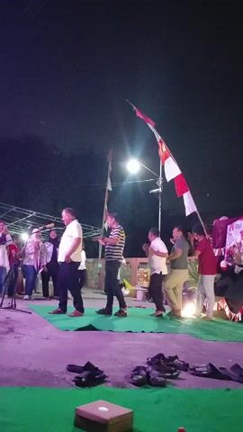The video captures an outdoor nighttime gathering. A group of people are standing on a raised platform covered with green fabric, with a purple-lit area in the foreground where shoes are placed. In the background, a stage is set up with lighting and a prominent Indonesian flag on a tall pole. Several individuals are on the stage, some appearing to be speaking or performing. The overall atmosphere suggests a community event or ceremony.