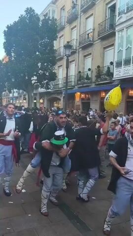 A man wearing a green cap and a white shirt is being carried on the shoulders of another man in a crowded outdoor street scene. The man on top is wearing sunglasses and a dark vest. Both men are wearing traditional Basque clothing, including white stockings with red ties. In the background, other people are dressed in similar attire, with women in red skirts and white blouses. A yellow Spongebob Squarepants balloon is visible in the upper right corner. The environment appears to be a festive gathering with buildings and trees lining the street.