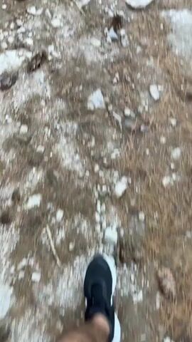 The video shows a first-person perspective of someone walking on a rocky, unpaved path through a forest. The path is lined with grass and trees. In the distance, there are a few people walking ahead on the path. The terrain appears to be slightly uneven with rocks and dry leaves scattered across the ground. The sky is overcast.