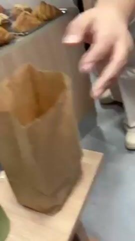 The video shows a bakery or cafe interior. Initially, a brown paper bag on a small wooden table is shown next to a green smoothie. The bag is then revealed to contain baked goods, including what appears to be a croissant. The camera pans to the right, showing a male barista wearing a light brown apron preparing a drink behind a wooden counter. There is a glass display case to his left with croissants in it. Other people can be seen moving around in the background.