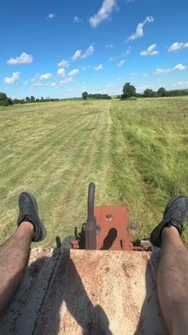 The video is a first-person perspective shot from the back of a red tractor. The tractor is in motion, cutting tall green grass in a wide field under a bright blue sky with scattered white clouds. The camera angle is low, showing the operator's legs and feet in black shoes. The tractor has a rusty metal deck and a single lever mechanism visible. The field is a mixture of cut and uncut grass, with rows of cut grass forming lines across the landscape. In the distance, trees and a slightly hilly horizon are visible. The overall scene depicts agricultural work on a sunny day.
