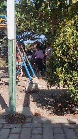 A group of people are gathered near a playground with swings and climbing structures. One person, wearing a light purple top and dark pants, is standing near a blue metal structure, looking away from the camera. To their right, a person wearing a white shirt and dark pants is also standing. Other people are visible in the background near other playground equipment. The area appears to be outdoors, with trees and sunlight casting shadows. The ground is covered in dirt and some paved areas.