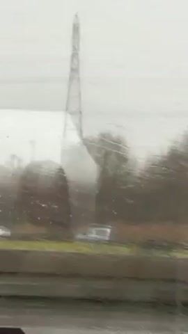 The video shows a view from inside a moving vehicle on a rainy day. Raindrops streak the window, partially obscuring the view outside. We see an electric pylon, trees, and a building with an arched doorway.  Cars are visible on the road. The scene is grey and overcast. The camera is stable, indicating a smooth ride.