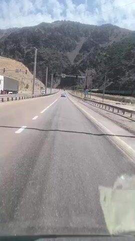 The video is a first-person perspective shot from inside a vehicle traveling on a road. The road has multiple lanes and is bordered by a metal guardrail. In the background, there are steep, rocky mountains with sparse vegetation. A train is visible on a track parallel to the road, moving in the same direction as the vehicle. Traffic lights and road signs are also present. The sky is blue with some clouds. The video appears to be filmed during the day, with bright sunlight.
