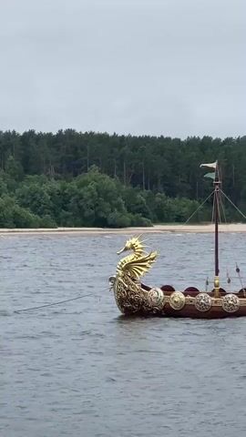 A medium shot shows a Viking-style boat with a dragon's head figurehead on a body of water. The boat is wooden with ornate carvings and shields along its sides. There is a mast with two flags and rigging. A person is visible in the boat near the stern, appearing to operate a small motor attached to the back. The water is choppy, and a line extends from the boat towards the left of the frame. In the background, there is a sandy beach and a dense forest. The sky is overcast. The camera is positioned at a slight angle to the boat, with a rocky foreground element on the right side.