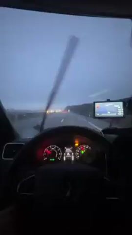 The view from inside a car shows a driver traveling down a wet road during dusk. The windshield wipers are on intermittently, suggesting light rain. The dashboard gauges are visible, and a GPS navigation system is mounted near the top of the dashboard, displaying a map. The car is a Renault, as seen by the logo on the steering wheel. There are other vehicles visible on the road ahead, indicated by their tail lights. The overall scene suggests a commute or trip during inclement weather.