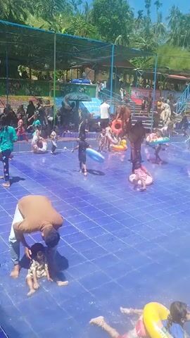 The scene depicts an outdoor splash pad area teeming with people, primarily children, enjoying the water on a sunny day. The splash pad is surfaced with square blue tiles. Many individuals are wearing swimwear, some are using inflatable rings, while others are walking or playing directly in the water. A man is bending down to assist a small child in the water. A few people are positioned along the perimeter, observing the activities. The setting is surrounded by greenery, with a few structures that appear to be part of the water park amenities. The atmosphere seems lively and joyful.