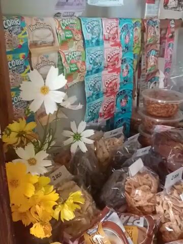 The video showcases a view of a small shop or kiosk displaying various products. The foreground features artificial daisies and yellow flowers arranged for decoration. Behind these, a shelf presents an assortment of items, including wrapped snacks, pastries, and containers of what appears to be dried food. There are some patterned scarves or cloths stacked to the left side. The composition is somewhat cluttered, providing a general sense of the shop's inventory and decorative style.