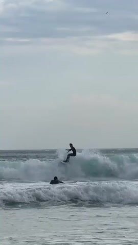 The video shows two people in the ocean on surfboards. One person is standing on their board and riding a wave. The other person is sitting on their board in the water. The ocean is choppy with white foamy waves. The sky is overcast.