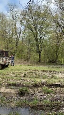 A blue and beige Nissan Xterra SUV is featured off-roading through a muddy, water-logged track in a wooded area. The vehicle, covered in mud, drives into and out of deep puddles, splashing mud and water high into the air, particularly from the front wheels. The setting appears to be a forest or grove with numerous trees displaying fresh, light green foliage, indicating springtime or early summer. The ground is a mix of mud, grass, and fallen leaves, with clearly defined tracks from previous vehicles. The overall scene suggests an adventurous off-road excursion in Manavi, Georgia, during daylight hours. No people are explicitly visible, but the presence of a driver is implied.