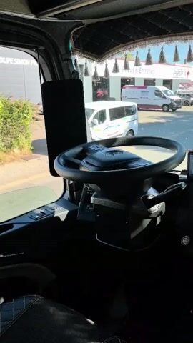 The video captures the interior of a truck cabin, looking out through the windshield. The focus is on the dashboard, prominently featuring a steering wheel with a Mercedes-Benz logo. Multiple screens and devices are mounted on the dashboard, including what appears to be a GPS unit and a large touchscreen infotainment system. Decorative elements, such as a patterned trim with tassels along the top of the windshield and what looks like prayer beads hanging from the gear shift, suggest a personalized interior. Outside the truck, a parking lot or service area is visible with several white vans and cars, some with company logos. The scene is brightly lit by daylight.