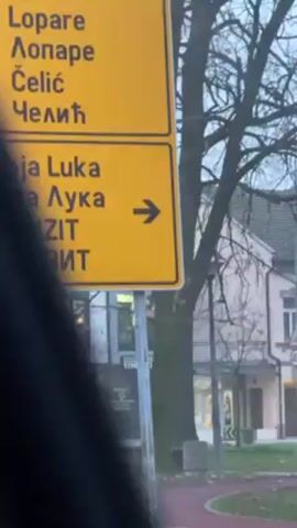The camera is inside a vehicle, capturing video of traffic and scenery. The vehicle is in an urban area, as indicated by the presence of buildings, trees, and street signs. Initially, the focus is on a yellow road sign listing place names in both Latin and Cyrillic script. The sign points towards Banja Luka. As the car moves, it passes a white flatbed truck adorned with graphics. It then focuses on another yellow sign that also lists Tuzla. The general atmosphere seems to be a quiet, slightly overcast day.