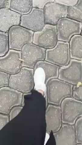 The video shows a person walking on a paved walkway. The paving consists of interlocking gray stones. The person is wearing white sneakers and long black pants. The camera is positioned above, looking down at the person's feet and the path. The person takes several steps forward. Some fallen leaves are visible on the pavement.