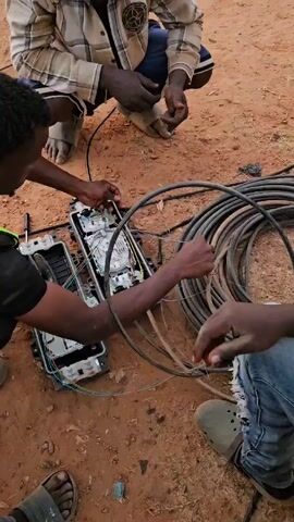 Three men are working on fiber optic cables. They are outside, kneeling or sitting on the ground, which appears to be a reddish-brown dirt. In front of them are two open black boxes containing wires and other components. A large coil of black cable lies nearby. The men are dark-skinned and are wearing casual clothing, including jeans, sandals, and plaid shirts. One of the men is looking at his phone, which displays an app with a red icon. They appear to be connecting or repairing the fiber optic cables.