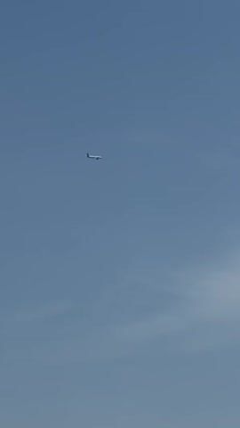 A white airplane is seen flying across a clear, bright blue sky during the daytime. The plane moves steadily from the left side of the frame towards the right, appearing as a small object high above. A faint, wispy cloud formation is visible in the lower right portion of the sky. No other objects, people, or discernible text are present in the video.