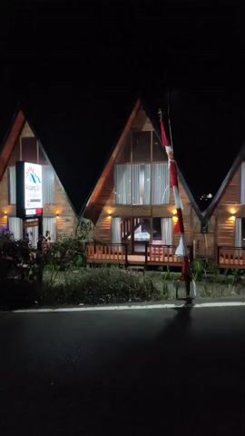 The video captures a nighttime scene of a street lined with modern, triangular-shaped buildings. The structures are made of wood and are illuminated by warm, ambient lighting, creating a cozy atmosphere. A prominent Indonesian flag is visible, standing tall and adding a splash of color to the dark surroundings. Several cars are parked along the street, suggesting a residential or commercial area. The video pans from right to left, revealing different angles of the buildings and the street. The overall impression is one of a tranquil and well-lit neighborhood, likely a resort or a collection of vacation homes.