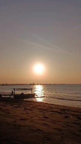 A vertical shot shows a serene beach scene at sunset. The sun, a bright orb, is positioned high in the sky, casting a warm golden glow and reflecting shimmering light across the calm ocean waves. The sky transitions from a pale blue at the top to a soft orange hue near the horizon. In the foreground, a sandy beach slopes gently towards the water. A small, dark-colored boat rests on the sand near the water's edge. A single figure is seated on the boat, facing the water. In the distance, several piers or structures extend out into the sea, silhouetted against the sunset. The overall atmosphere is tranquil and picturesque.