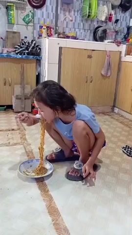 In a kitchen, a young girl crouches to eat noodles from a plate on the floor. The girl has dark hair and wears a light blue shirt and printed shorts, and has gray, furry sandals on her feet. She uses a spoon to lift long strands of noodles to her mouth. The kitchen has tiled floors and light wooden cabinets. A sink with hanging dish-drying racks is visible behind her.