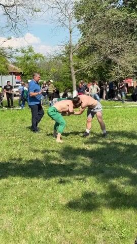 Two men are wrestling on a grassy field in an outdoor setting. One man is wearing green pants and is barefoot, while the other is wearing shorts and black shoes. A man in a blue shirt is standing between them, possibly acting as a referee. A crowd of people is watching the wrestling match in the background. The setting appears to be a park or field with trees and a blue sky. There is music playing in the background.