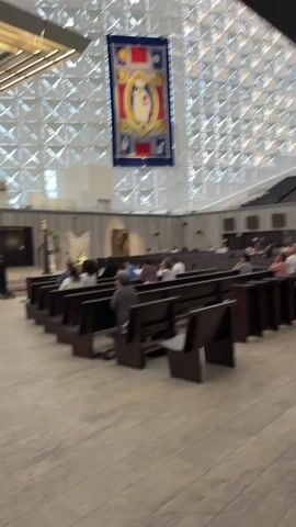The video shows the interior of a large, modern church with a geometric patterned ceiling. In the foreground, rows of dark wooden pews face an altar area. Several people are seated in the pews, facing the altar. Two individuals, dressed in dark robes, are kneeling at the altar. Above the altar, a large crucifix hangs from a modernist lighting fixture. To the left of the altar, a cross is visible. In the background, a large tapestry depicting a religious figure hangs from the ceiling. The environment appears to be dimly lit, with light filtering through the ceiling structure. Organ pipes are visible on a balcony in the background. The overall atmosphere suggests a religious service or ceremony.