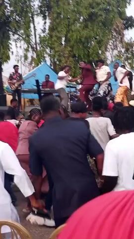The video shows an outdoor gathering with a live band performing on a stage. Several people are dancing and interacting with each other. A singer in a maroon suit is on stage, singing into a microphone, with other band members playing instruments like a guitar and keyboard. A crowd of people are gathered in front of the stage, some dancing energetically, others watching the performance. The environment appears to be a public space with trees and buildings in the background. The lighting suggests it is daytime.