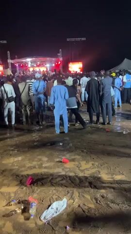 A nighttime outdoor concert with a crowd of people facing a stage with bright lights and video screens. The ground is muddy and littered with plastic cups and bottles. Many people are wearing traditional Nigerian clothing. The crowd is lively, with some people raising their hands.