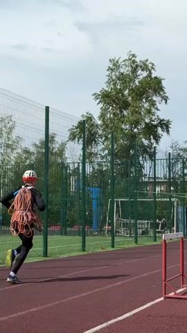 A person in black leggings and shirt, yellow athletic shoes, and a white helmet, runs on a red running track carrying a large orange rope coil. The track is adjacent to a green chain-link fence. There are several small red and white hurdles on the track. Beyond the fence are various objects including a soccer goal, a basketball net, and other unidentifiable items. To the right of the track is a large green tent. Trees are scattered around the area and the sky is cloudy. The person is running away from the camera, towards the tent.