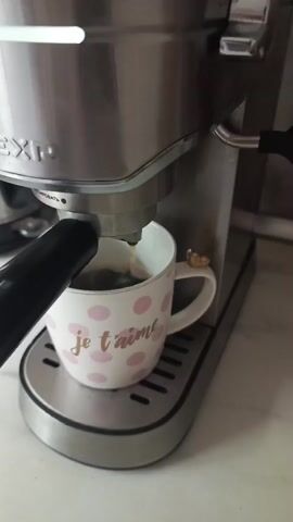 The video shows an espresso machine brewing coffee into a mug. The mug is white with pink polka dots and the words "je t'aime" in gold lettering. A small golden charm shaped like a heart is attached to the handle of the mug. The coffee is dark brown and is seen dripping from the spout of the machine into the mug, creating a crema on top. The machine is made of stainless steel and has a black handle. The background appears to be a kitchen counter, with a tiled wall visible behind the machine. The lighting is bright, suggesting it is daytime.