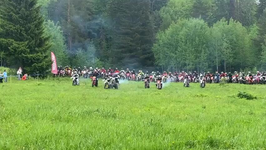 The video depicts a group of people participating in a motocross event. The racers, wearing helmets and protective gear, are starting their motorcycles on a grassy field.  They are surrounded by a backdrop of lush green trees and foliage. Spectators can be seen standing behind a blue rope, observing and taking pictures.  A red flag is visible in the distance. The scene has a smoky haze, suggesting the bikes have just started up. The camera focuses on the racers in the foreground as they begin to move.