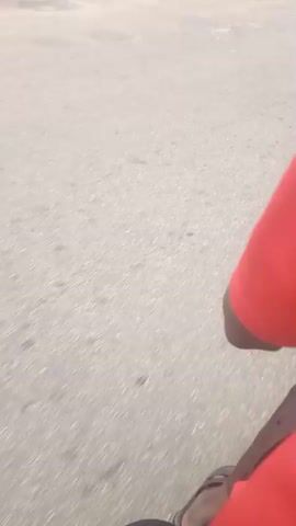 The video shows a person in a red shirt walking in an outdoor environment. The person is moving forward and looking towards the right. In the background, there are trees, a road, a sign with some writing, and a monument with insignia. The sky is cloudy. The camera is tilted and is moving, suggesting it is being held by the person. The scene appears to be a campus or an institutional area.