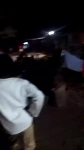 The video is taken at night and is mostly dark, with a few overhead lights in the distance.  A person wearing a white long-sleeved shirt is walking while recording the video. There are other people present, and what appears to be red and blue chairs or containers are visible in the background. There's indistinct background noise, possibly music and speech, in the background.