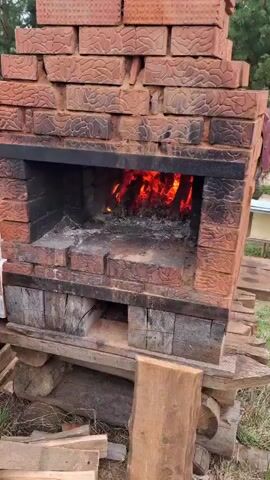 The video shows a brick oven with a fire burning inside. The oven is constructed from reddish-brown bricks with a textured pattern and reinforced with black metal supports. The interior of the oven is lined with similar bricks, now coated with ash and soot.  A fire is blazing inside, with glowing embers and flames visible. The oven is elevated on a wooden platform made of rough-cut planks.  The surrounding environment appears to be an outdoor setting, with trees and grass visible in the background.