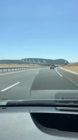 The video shows a point-of-view shot from inside a vehicle traveling on a highway. The camera is looking forward through the windshield. The highway has multiple lanes separated by white lines and a guardrail on the left. In the distance, a large, modern bridge with distinctive V-shaped pillars is visible spanning across a valley or body of water, with a hill on the far side. Several other vehicles are on the highway ahead, appearing to be moving in the same direction. The sky is clear and blue, indicating a sunny day. The dashboard of the vehicle is visible in the foreground.