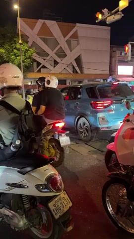 The video captures a street scene at night, likely at an intersection or busy road. Several motorbikes and a blue SUV are visible, suggesting traffic. The environment appears to be urban, with buildings and streetlights illuminated. One segment shows a gas station with a woman in a pink helmet on a motorbike, followed by a white tuk-tuk driving past. The camera is mostly focused on the motorbikes and the general surroundings, with occasional glimpses of people on the vehicles.