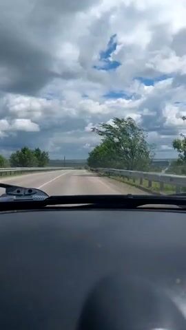 The video is filmed from the interior of a vehicle, looking out through the windshield. The road ahead is a two-lane asphalt road that curves to the left. Trees line the right side of the road, with metal guardrails running alongside them. The sky is filled with dramatic, fluffy white clouds interspersed with patches of blue. The landscape on either side of the road appears to be rural, with fields and distant hills. The wind is visibly strong, causing the trees on the right to sway dramatically. A road sign is visible on the right side of the road. The dashboard of the car is visible at the bottom of the frame.