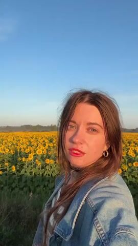 A young woman with long brown hair and blue eyes, wearing a denim jacket over a white shirt and a black choker necklace, stands in a field of sunflowers. Her lips are painted red. The sun is setting, casting a warm glow and shadows across her face and the field. The sky is a clear blue with a few wispy clouds. She is looking at the camera and smiling. The video is a selfie, as indicated by the camera angle.