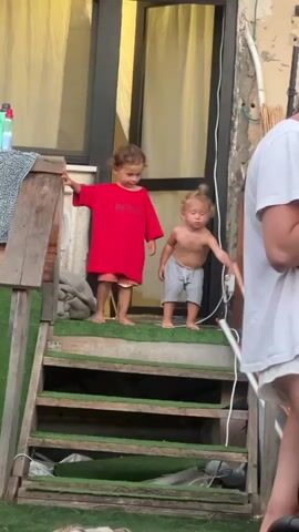 Two young children are on a set of wooden outdoor stairs, each covered with artificial turf. The child on the left wears a red t-shirt and orange shorts, while the child on the right is shirtless and wears grey shorts. They are standing on the top step. The child on the left leans against a wooden railing and appears to be looking down. The child on the right is looking towards the child on the left and seems to be interacting with them. The background shows a glass door with yellow curtains, and a textured wall. A person in a white t-shirt is partially visible on the right side of the frame. The scene appears to be outdoors, possibly in a backyard or patio area, and the lighting suggests it is daytime. The overall atmosphere seems casual and familial. There is text visible on the red shirt of the child on the left which reads "BOSS". The image has been captured in Kiryat Yam, Israel.